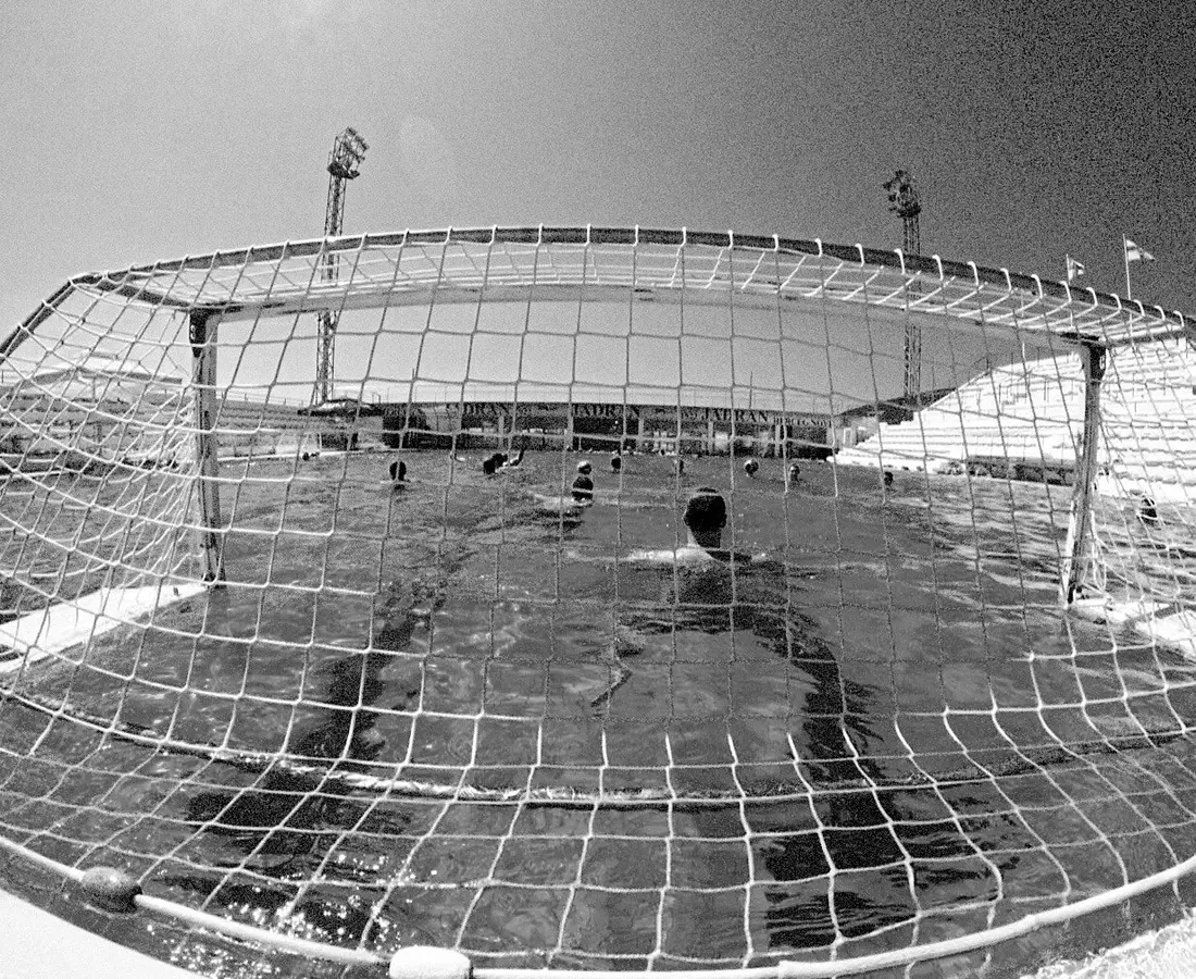 A water polo goal and net seen from the pool edge, with a player in the water